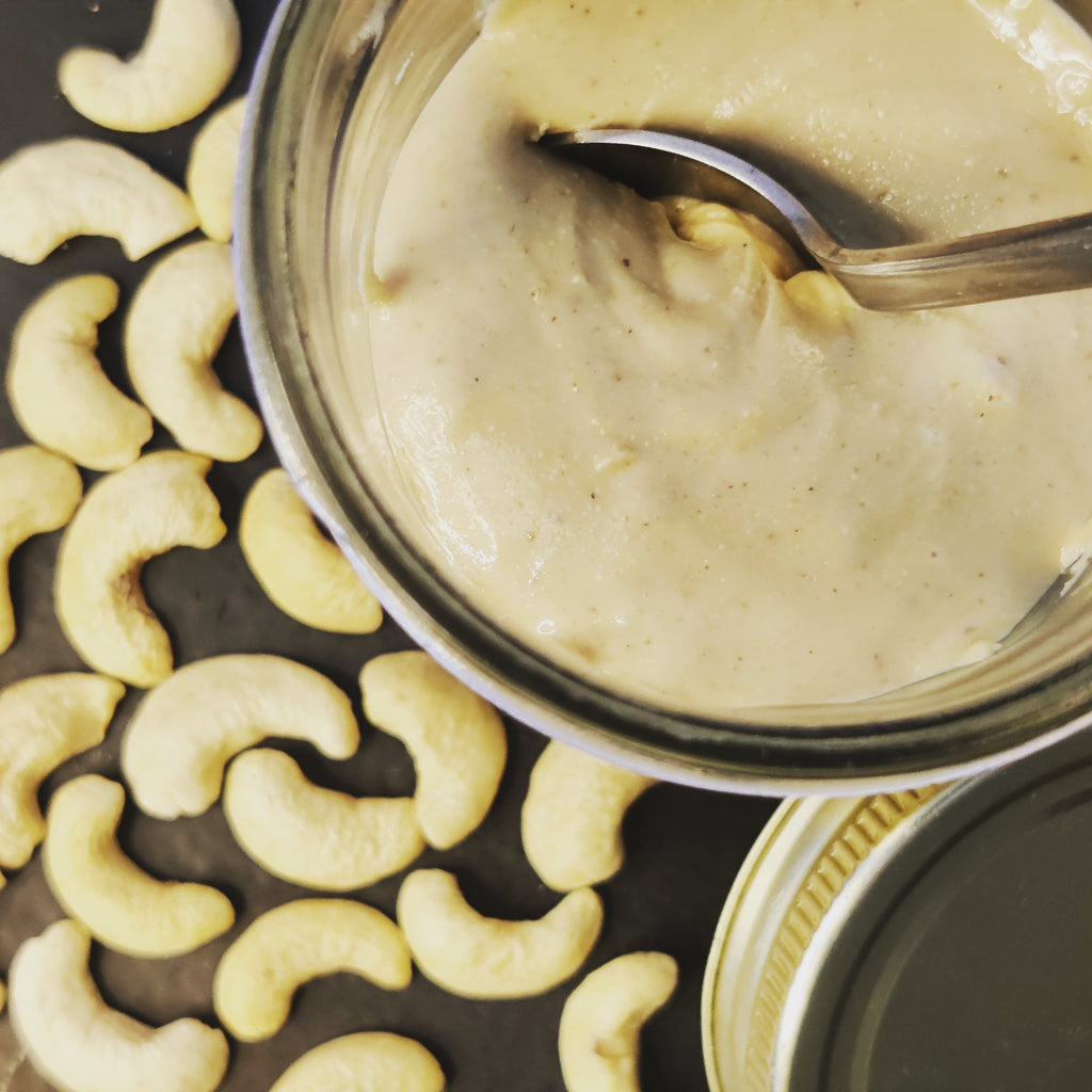 Jar of creamy spread with a spoon, surrounded by crescent-shaped cashews on a dark surface. Premium cashew butter
