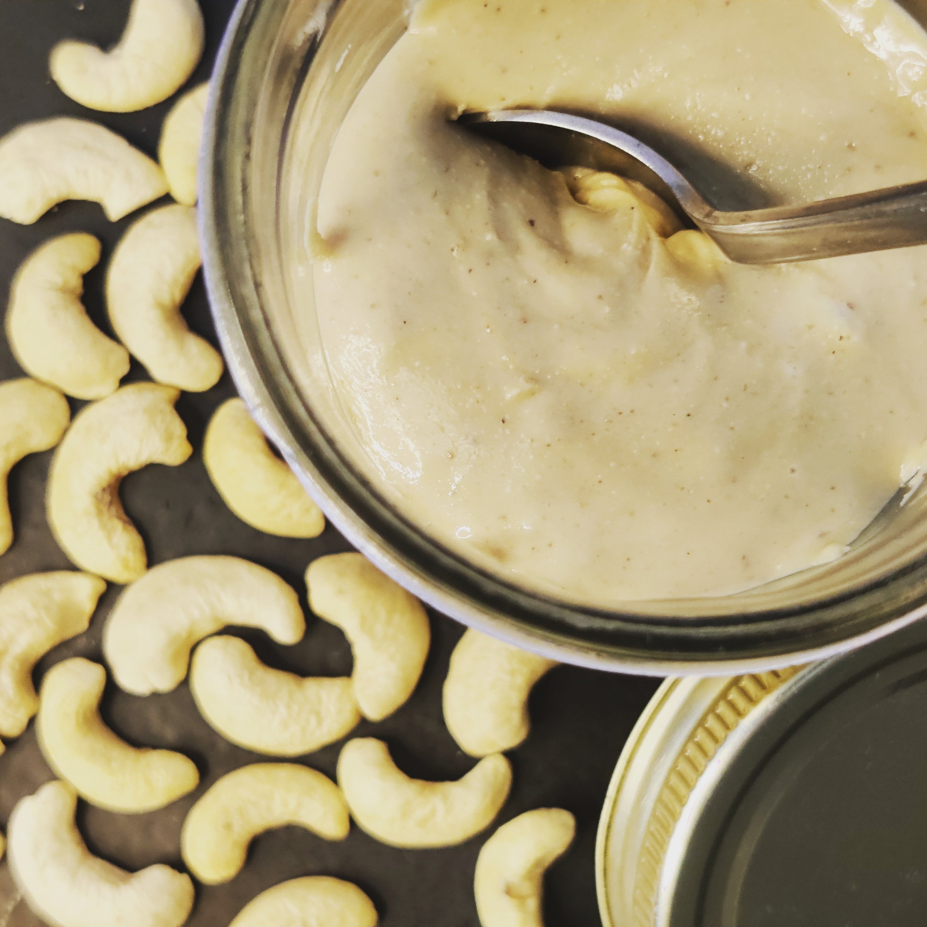 Jar of creamy spread with a spoon, surrounded by crescent-shaped cashews on a dark surface. Premium cashew butter