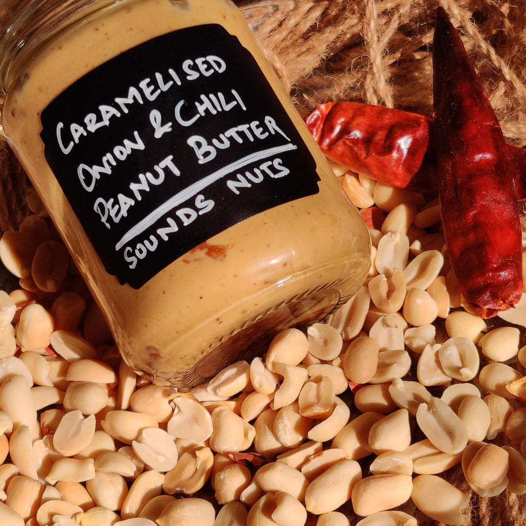 Jar of caramelized onion and chili peanut butter surrounded by peanuts and red peppers on a woven surface.