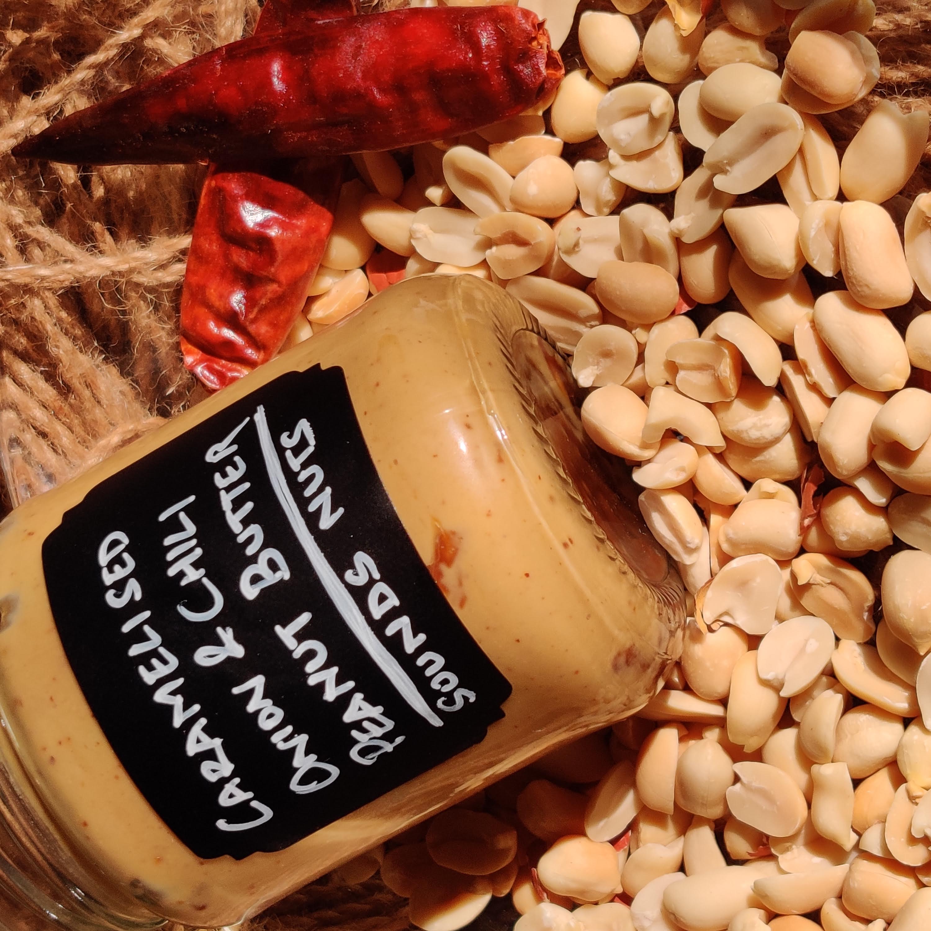Jar of caramelized onion and chili peanut butter surrounded by peanuts and red peppers on a woven surface.